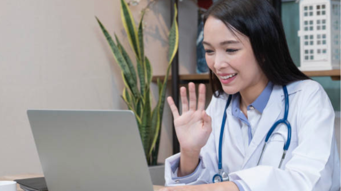 A female medical professional of Asian ethnicity in a white coat waves to a laptop, ready for her free coaching consultation.