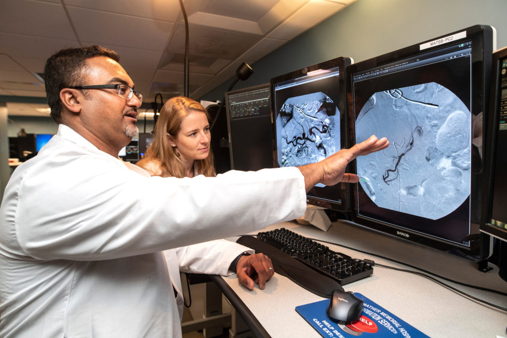 A male radiologist wearing glasses and a white coat points at a computer screen displaying medical images. A female radiologist with shoulder-length hair leans in to view the screen, both engaged in professional discussion in a medical imaging room.